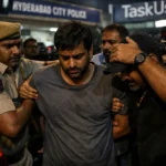 Individual in distress being escorted by police under bright lights.